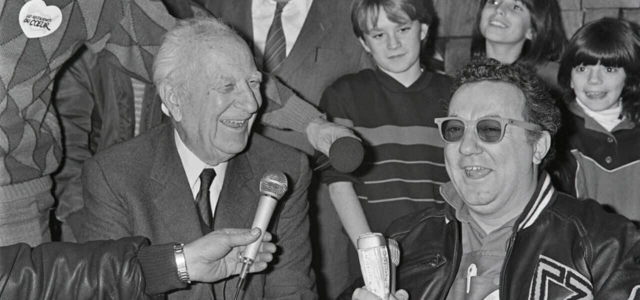 Coluche et le maire de Marseille Gaston Deferre présentent à la presse le 18 janvier 1986 une campagne en faveur des Restos du Cœur et plaident pour l’élaboration d’une loi donnant plus de pouvoir aux associations caritatives. À l’occasion de cette visite, une opération de récupération de denrées alimentaires sera organisée sur les parkings de 14 supermarchés de la région. © Pierre Ciot / AFP